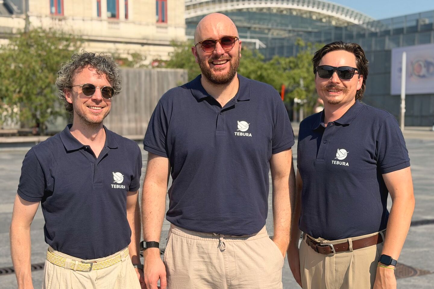 The three Tebura founders standing side by side in navy Tebura polo shirts on a sunny city square